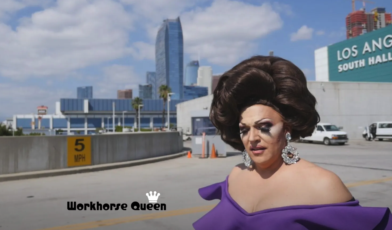 Drag queen in purple dress, large wig, and makeup, stands outdoors with Los Angeles buildings in background.